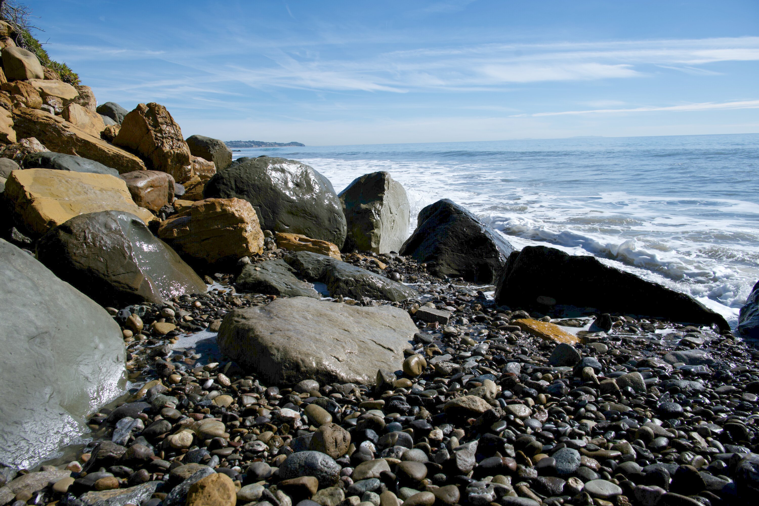 Golden and gray rocks scattered along Malibu Beach with gentle waves in the background on a sunny winter day.
