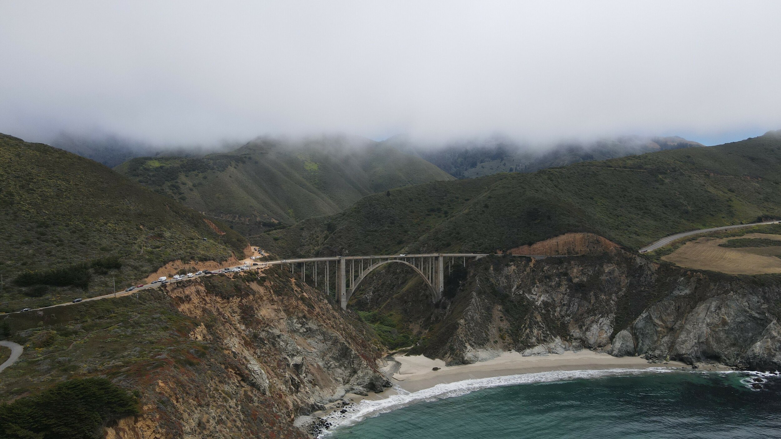 Aerial photograph of Bixby Bridge enveloped in fog, with the Pacific Ocean coastline and green mountains.